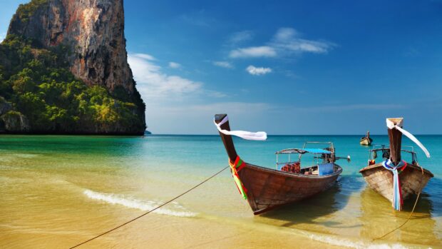 Traditional long tail boats moored on the sandy shore of Thailand with clear blue sea and limestone cliffs