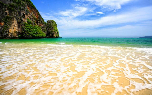 A scenic Thailand coastline with golden sand and lush green cliffs under a blue sky