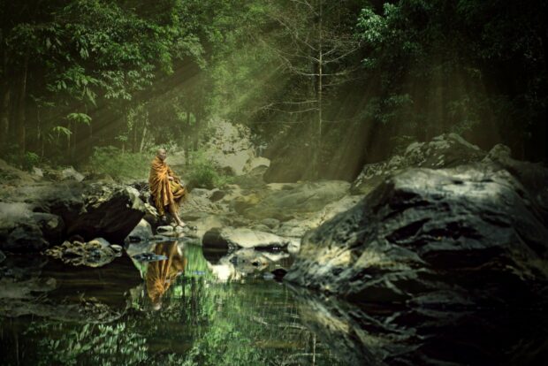 A monk in traditional robes meditating by a calm river in Thailand forest