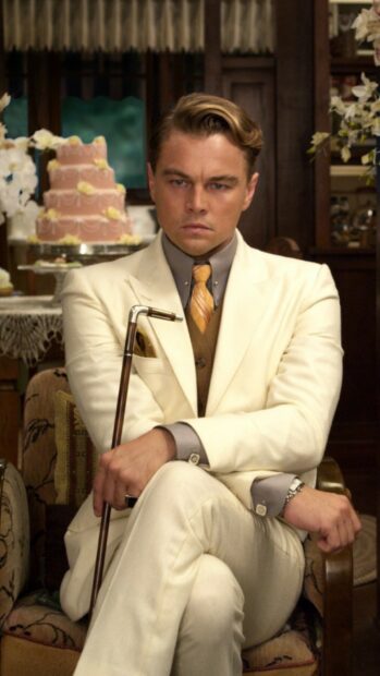 A man in a vintage suit holding a cane sitting in a classic room with a cake in the background