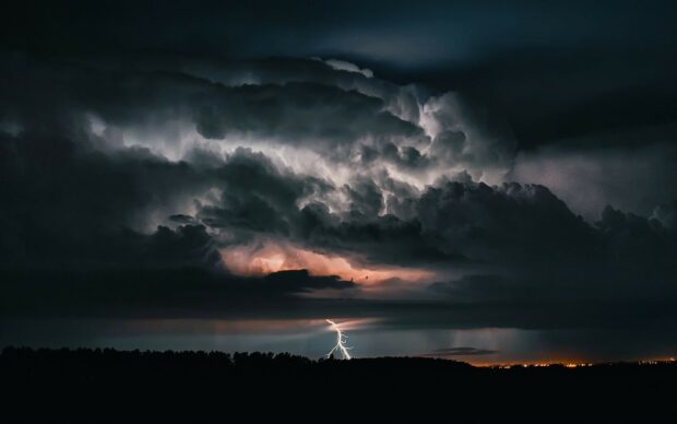 Dark storm clouds with bright thunder striking above forested horizon at night