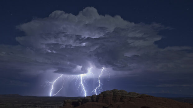 Thunder strikes illuminate the sky above rocky terrain during a nighttime storm