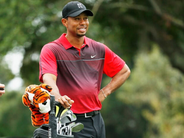 Tiger Woods standing with golf clubs and a tiger headcover on the golf course in a red shirt