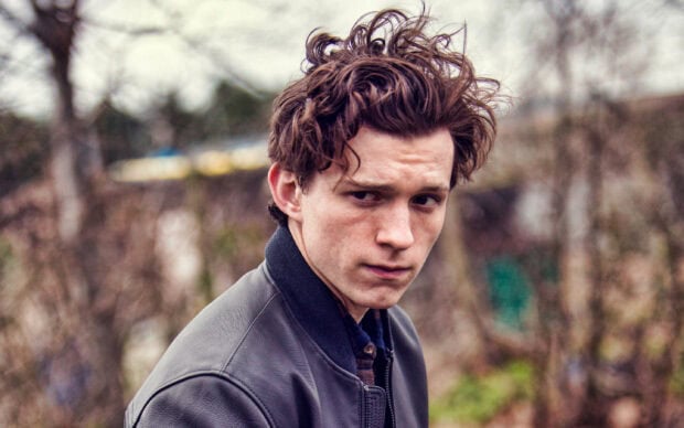 Tom Holland looking serious outdoors with messy hair wearing a leather jacket