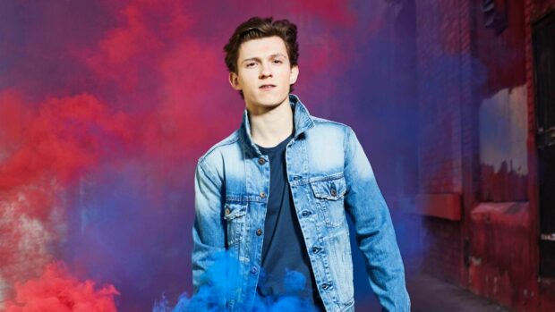 Tom Holland wearing a denim jacket posing with colorful smoke in the background