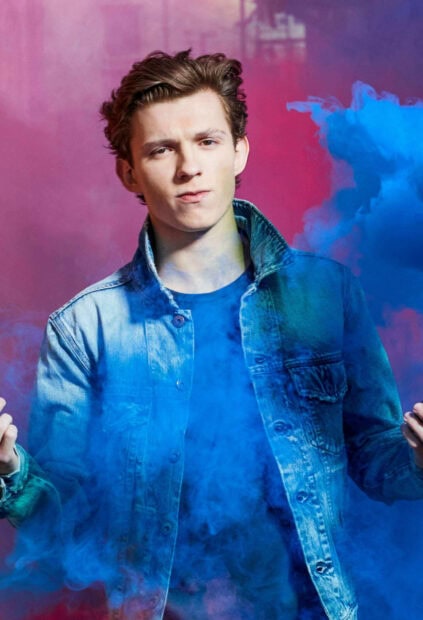 Tom Holland wearing a denim jacket surrounded by blue smoke against a red background