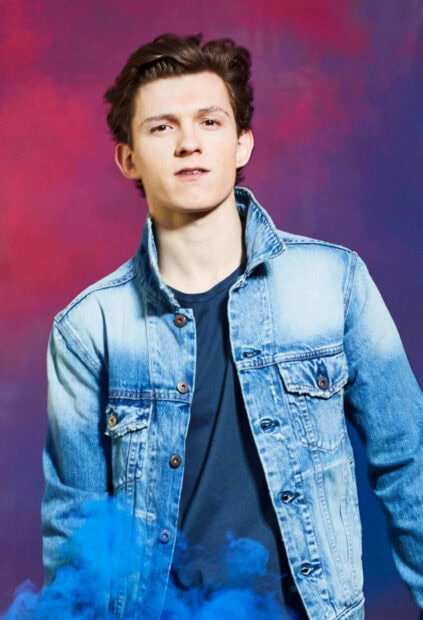 Tom Holland wearing a denim jacket with a colorful background and blue smoke