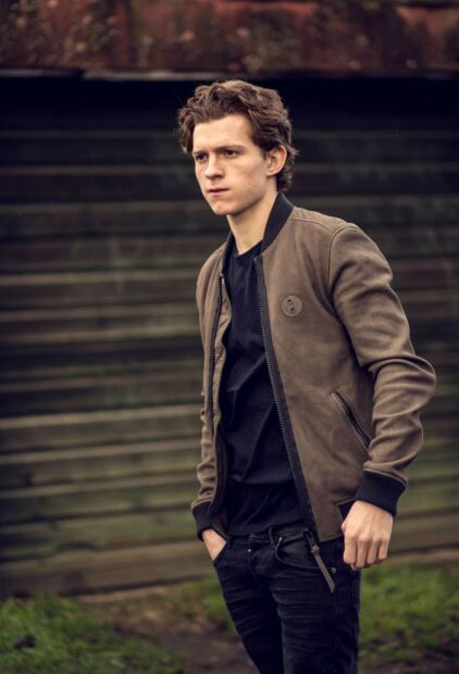Tom Holland wearing a brown jacket standing outdoors against wooden steps background