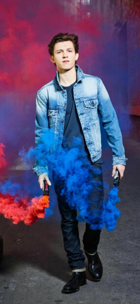 Tom Holland holding red and blue smoke bombs while wearing a denim jacket