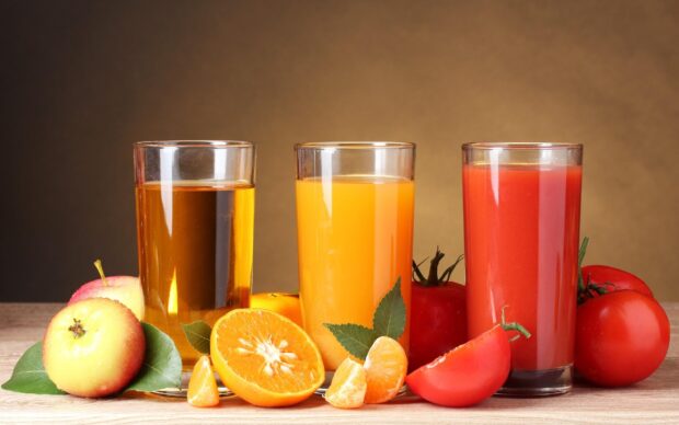 A glass of fresh tomato juice with whole and sliced tomatoes placed next to glasses of apple and orange juice