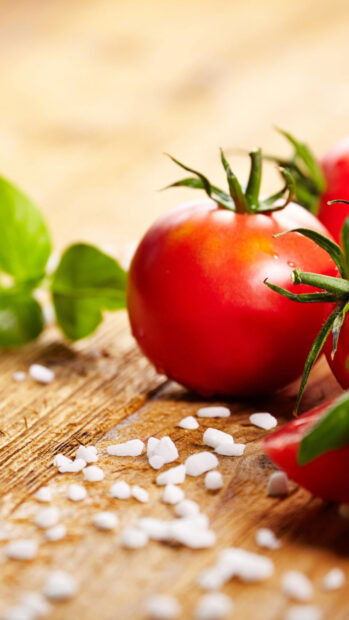 Fresh tomato with green leaves and coarse salt on wooden surface