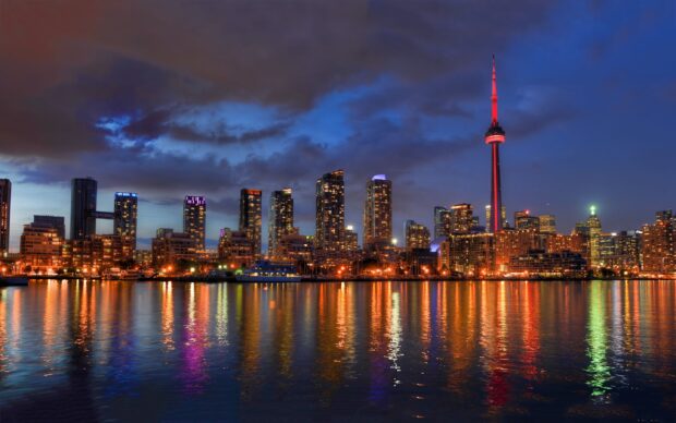 The Toronto skyline features illuminated skyscrapers and the CN Tower reflected in calm water at dusk