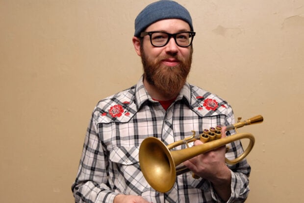 Man with beard holding trumpet wearing plaid shirt with floral embroidery