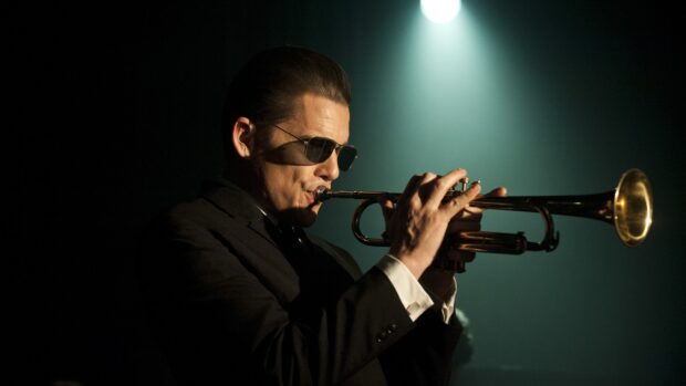 A man wearing sunglasses playing trumpet on stage with spotlight in background