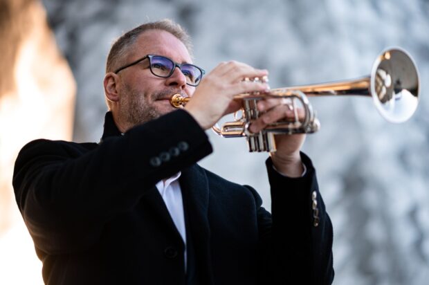 Man playing trumpet outdoors in a black coat and glasses