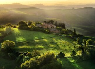 Rolling green hills in Tuscany with an old farmhouse surrounded by lush trees and sunlight filtering through the landscape