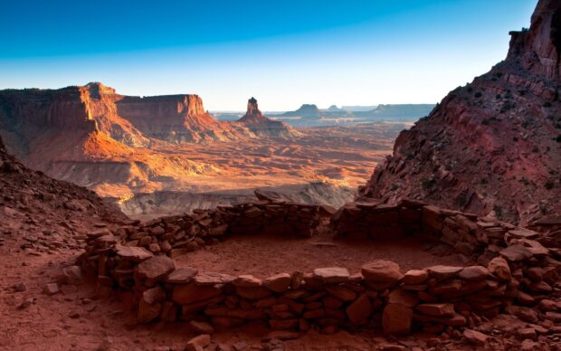 Ancient rock shelter ruins overlook a vast Utah desert landscape with red sandstone formations and clear sky