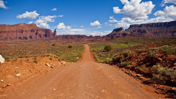 A scenic Utah desert landscape with a dirt road leading towards red rock formations under a blue sky with clouds