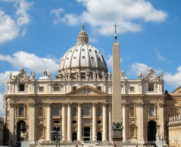 The dome of Vatican City Basilica with statues and an obelisk under a blue sky