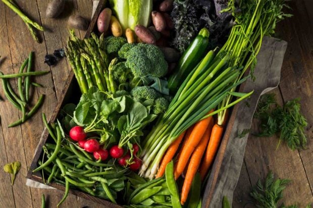 Fresh vegetables including carrots radishes broccoli and green beans arranged in a wooden tray on a rustic table