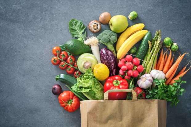 Fresh vegetables spilling out of a paper bag on a stone surface with avocado broccoli and carrots