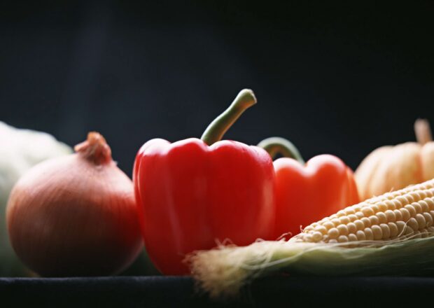 A group of fresh vegetables including red bell pepper onion and corn on a dark surface