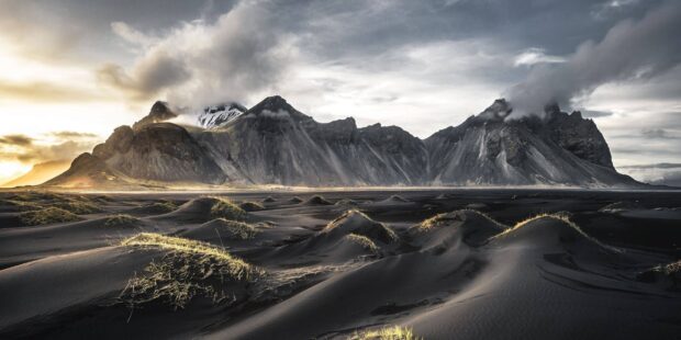 Black sand dunes with grass and rugged Vestrahorn Iceland mountains under cloudy sky