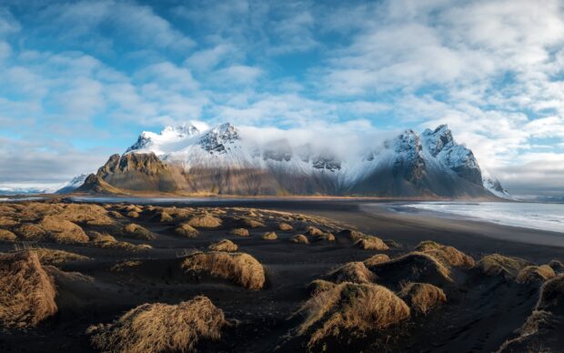 Snow capped peaks of Vestrahorn Iceland surrounded by black sand dunes and dry grass under a blue cloudy sky