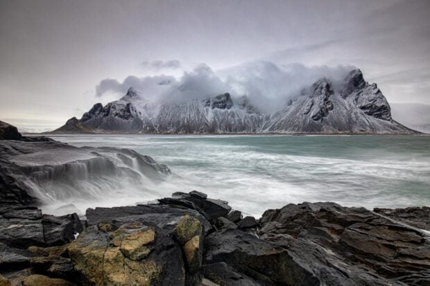 Dramatic rocky coast with Vestrahorn Iceland mountain covered by clouds and snow