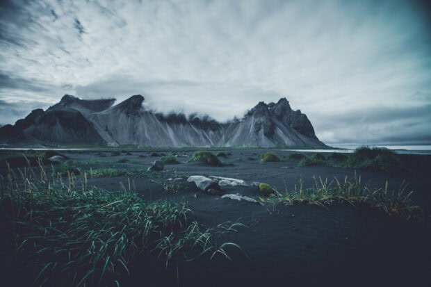 Black sand beach and rugged mountains of Vestrahorn Iceland with green grass under cloudy sky