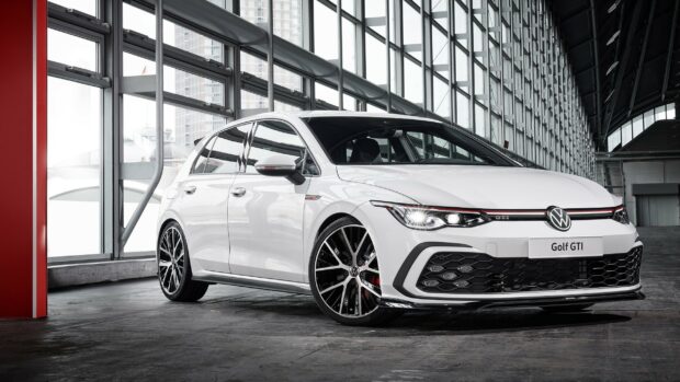 White Volkswagen Golf GTI parked in a modern glass building interior with sporty black wheels and headlights on
