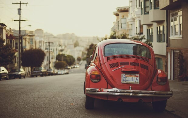 A red Volkswagen parked on a city street with buildings and trees in the background