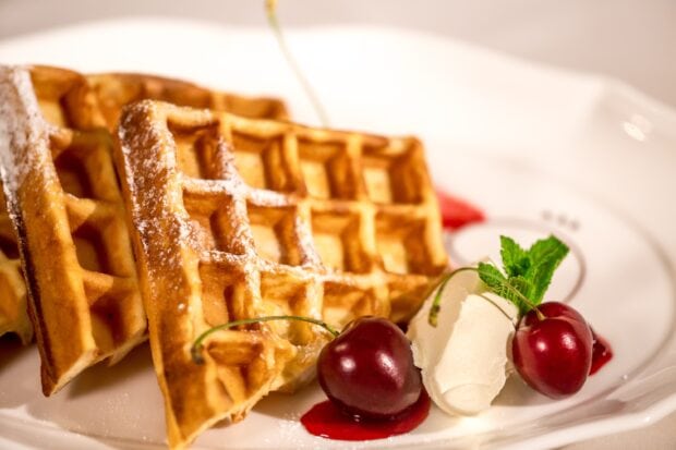 Golden waffle slices with cherries and cream served on a white plate with powdered sugar