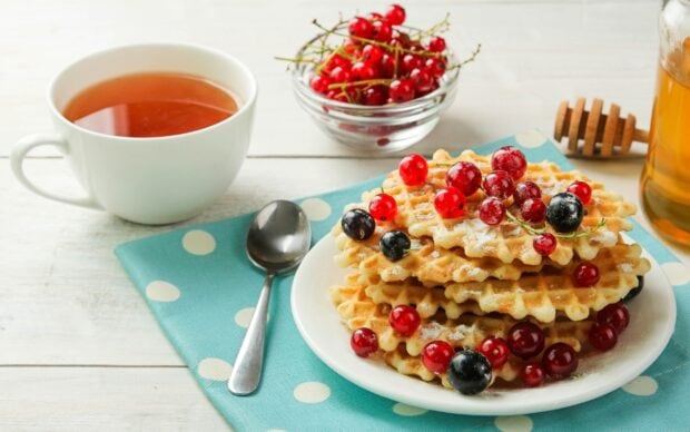 A stack of waffle topped with assorted berries on a plate next to a cup of tea and honey jar on a polka dot napkin
