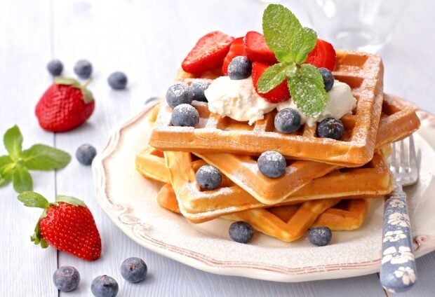 A stack of waffle topped with strawberries blueberries whipped cream and mint leaves on a ceramic plate