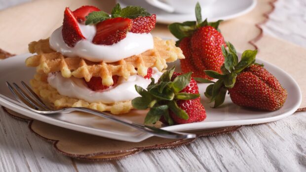 Fresh waffle topped with cream and sliced strawberries on a plate alongside whole strawberries