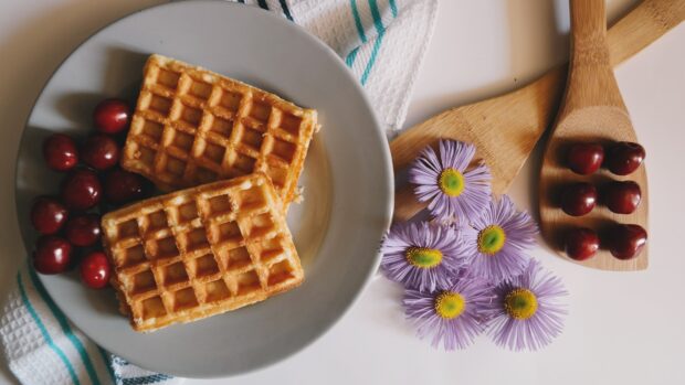 Two golden waffle pieces with cherries on a plate next to wooden spoons and purple flowers