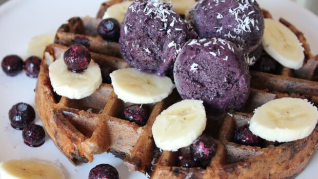 A close up of waffle topped with banana slices blueberry ice cream and fresh blueberries