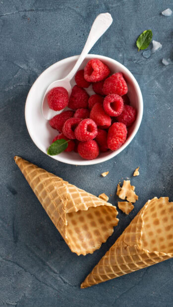 A bowl of fresh raspberries with a spoon next to two waffle cones on a textured surface