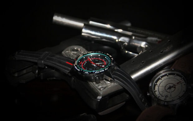 Black tactical watch with compass dial resting on a handgun in low light conditions