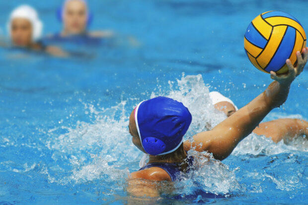 A water polo player wearing a blue cap prepares to throw the ball in the pool