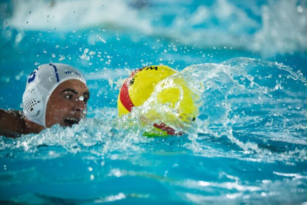 A water polo player wearing a white cap swimming towards the ball in a pool with splashing water