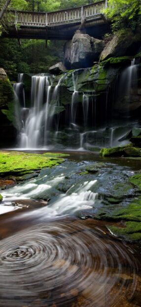 A beautiful West Virginia waterfall flowing under a wooden bridge surrounded by lush green moss and rocks