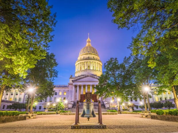 The West Virginia state capitol building with a historic bell in front surrounded by trees at dusk
