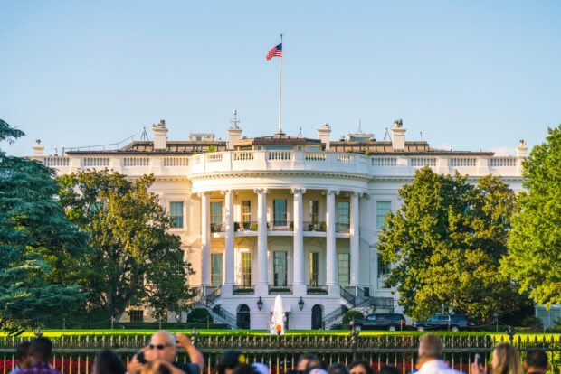 The White House building with the American flag and surrounding trees on a clear day