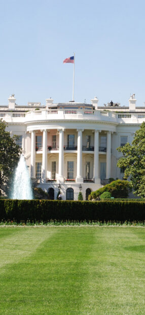 The iconic White House with the American flag flying above a clear blue sky and a green lawn in front