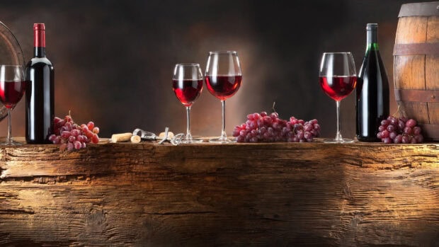 Red wineglass and grapes on a rustic wooden table with wine bottles and corkscrew