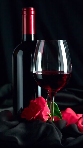 A wineglass filled with red wine next to a wine bottle and a red rose on black fabric