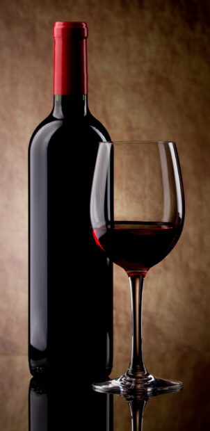 A wineglass filled with red wine in front of a wine bottle on a reflective surface