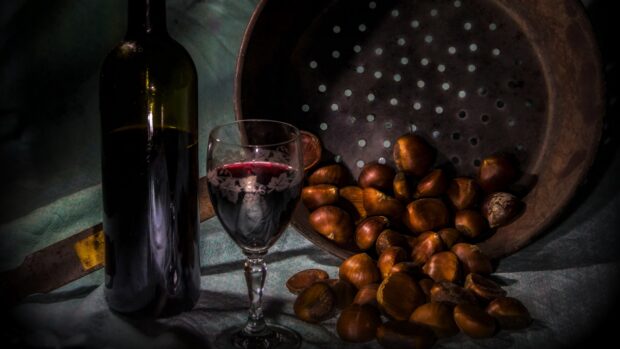 A wineglass filled with red wine beside a dark wine bottle and scattered chestnuts in a rustic setting